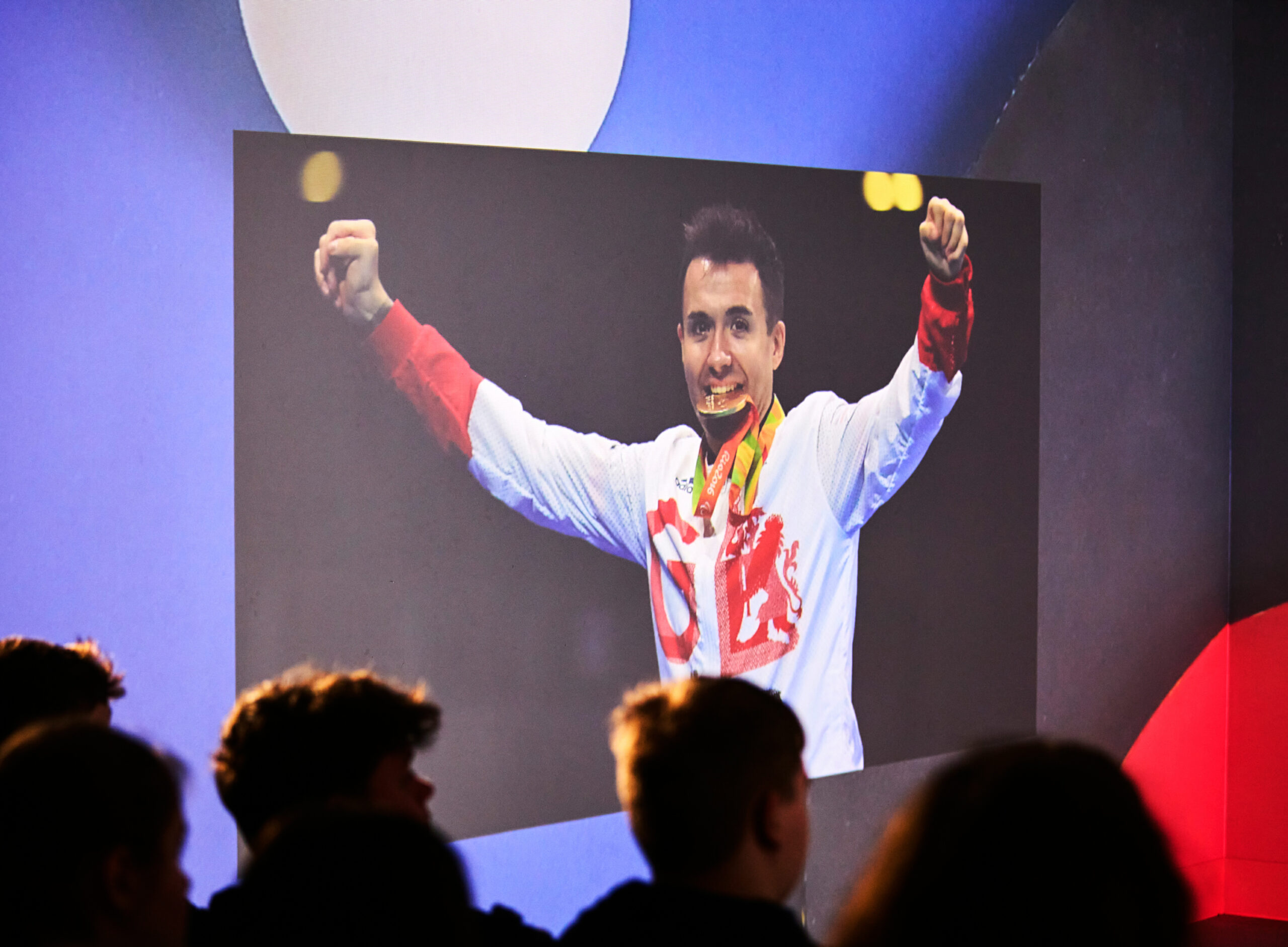 Three silhouetted heads look on at a projection of a male athlete in a GB training top, a gold medal clenched between his teeth and his arms raised in celebration.